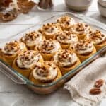 A glass baking dish of frosted Pumpkin Cinnamon Rolls topped with pecans.