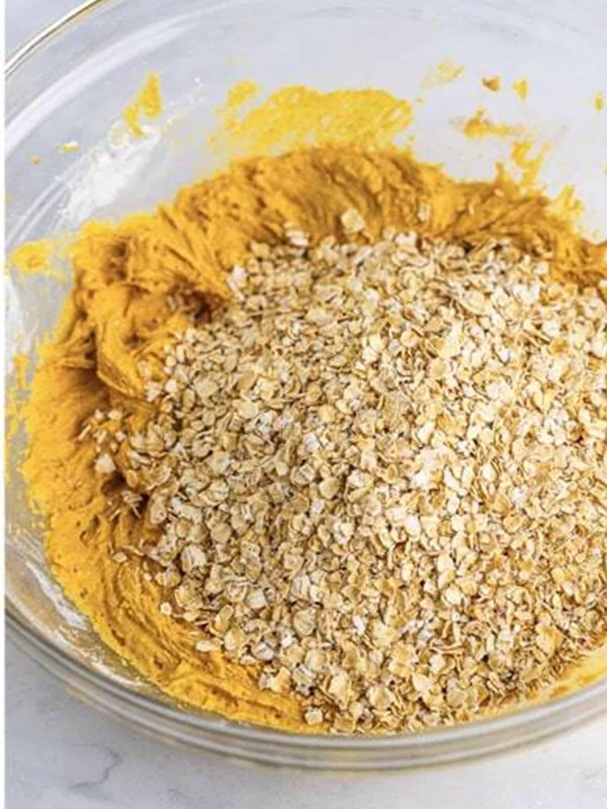 Rolled oats added to the thick pumpkin cookie dough in a glass mixing bowl.