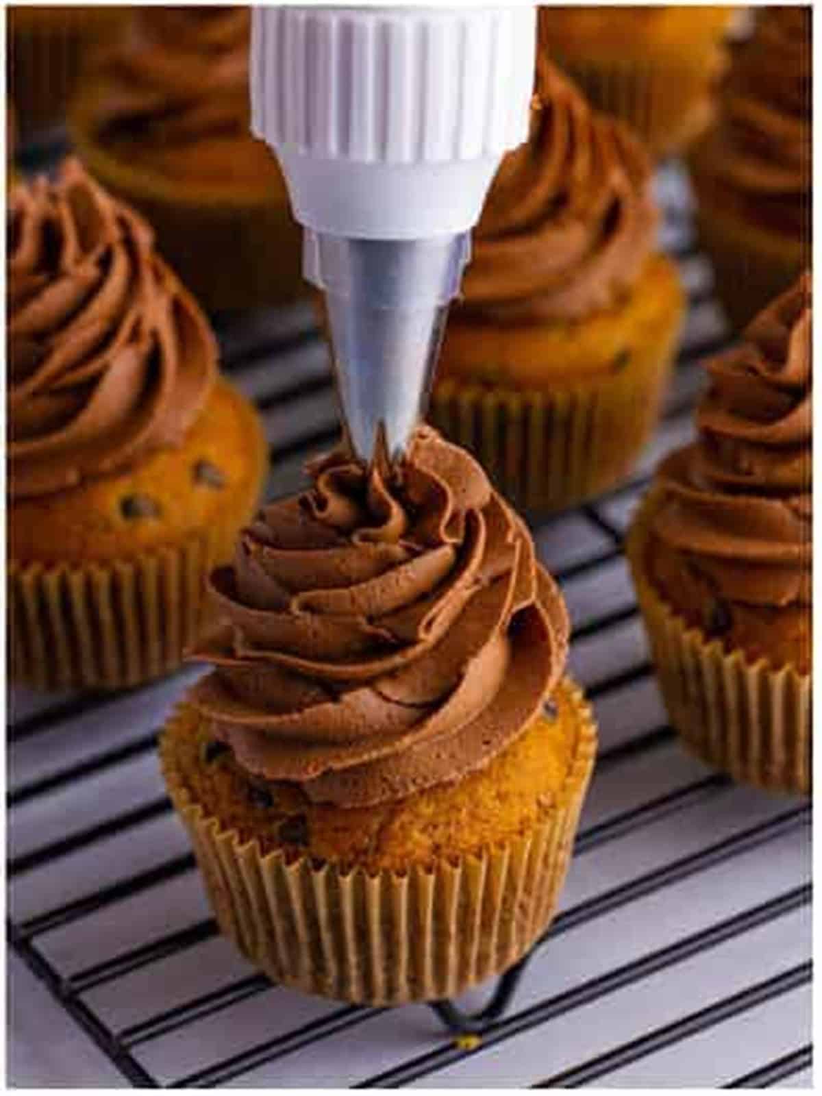 Piping swirls of chocolate frosting onto cooled pumpkin cupcakes on a wire rack.