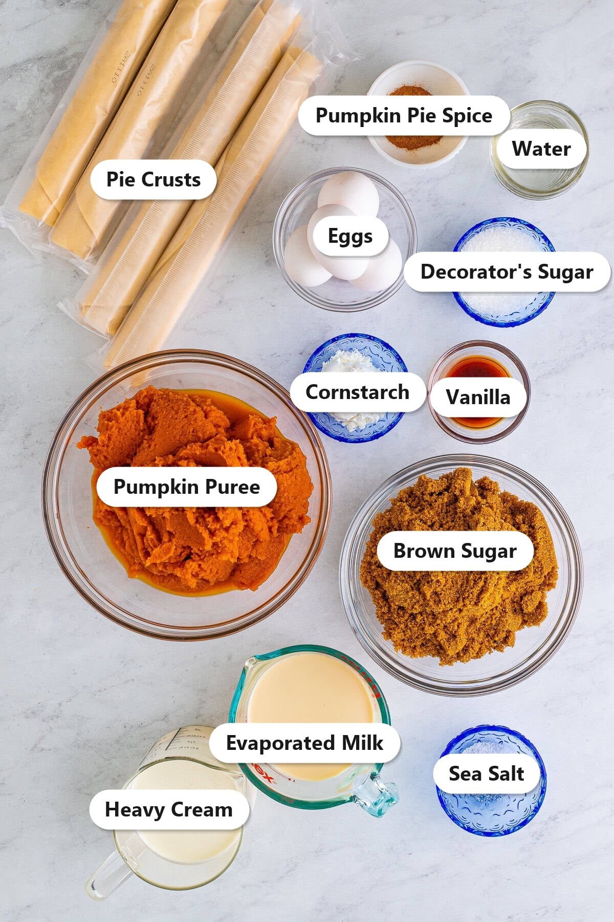Labeled ingredients for pumpkin slab pie: pie crusts, pumpkin puree, brown sugar, heavy cream, evaporated milk, eggs, vanilla, cornstarch, pumpkin pie spice, sea salt, water, and decorator sugar.