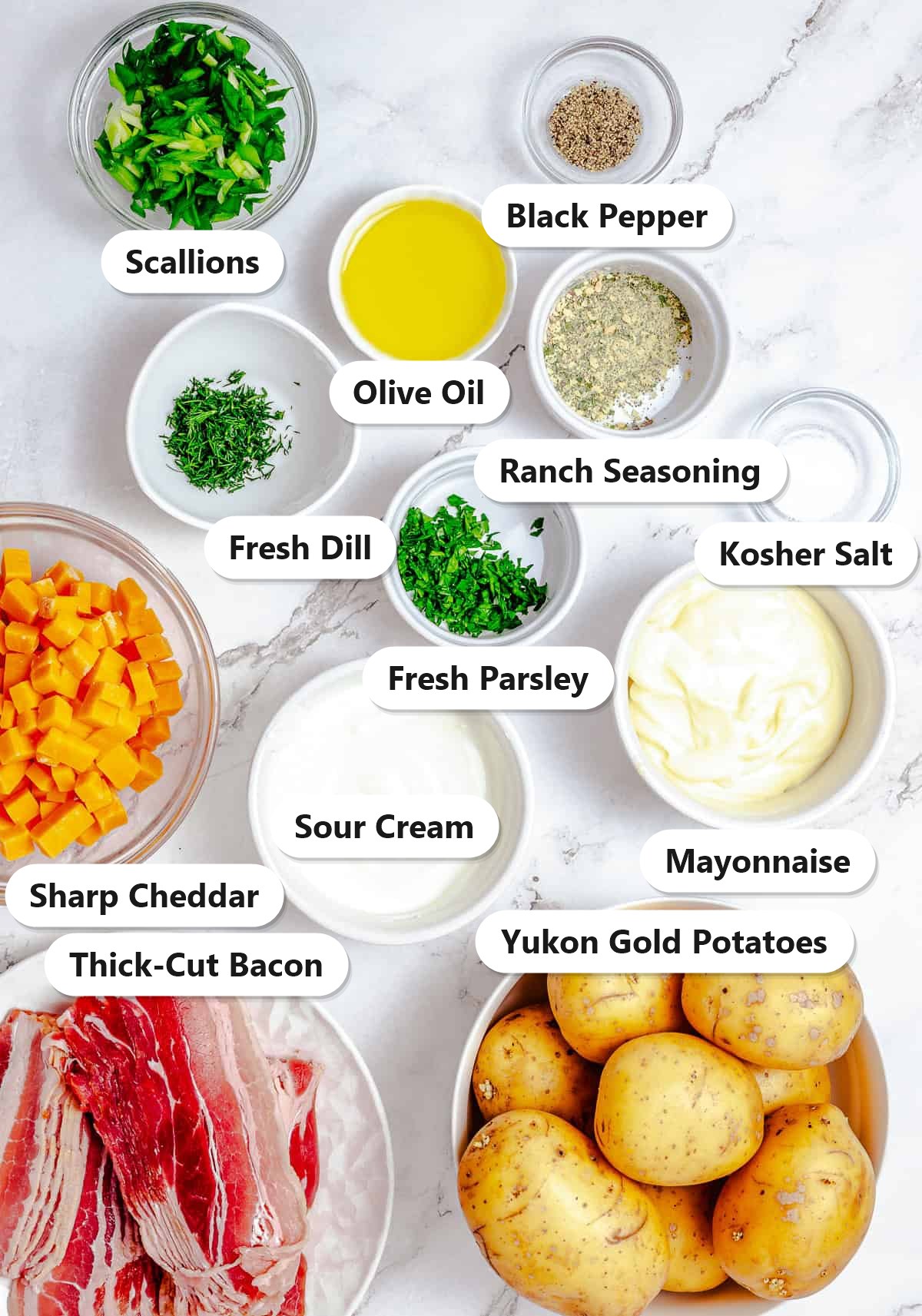 Labeled ingredients for Ranch Potato Salad including Yukon gold potatoes, thick cut bacon, sharp cheddar, mayonnaise, sour cream, ranch seasoning, dill, parsley, scallions, olive oil, salt, and pepper.