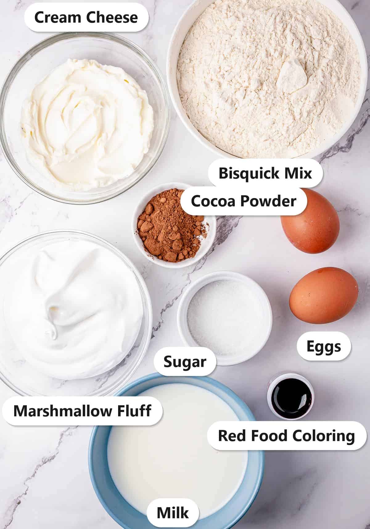 Ingredients to make red velvet pancakes labeled on a marble surface: cream cheese, Bisquick mix, cocoa powder, marshmallow fluff, sugar, eggs, milk, and red food coloring.