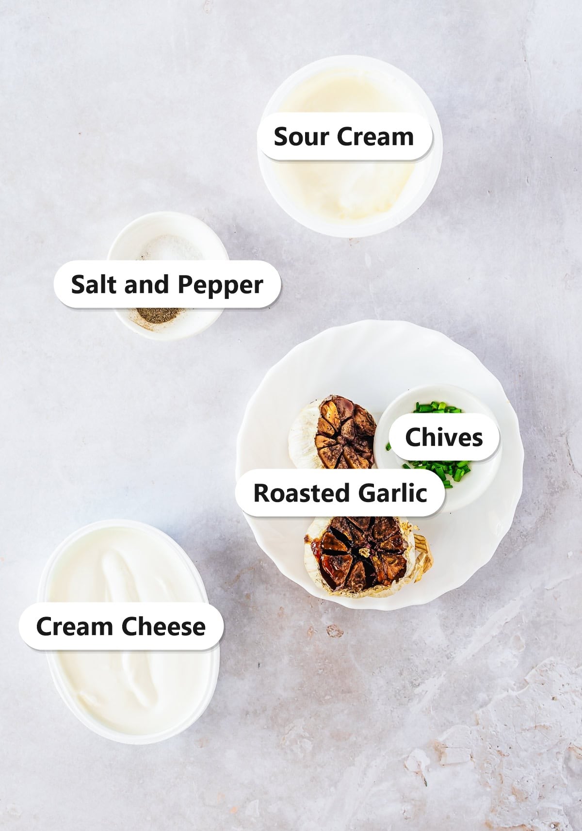 Labeled ingredients to make roasted garlic cream cheese dip including cream cheese, sour cream, roasted garlic, chives, and salt and pepper.