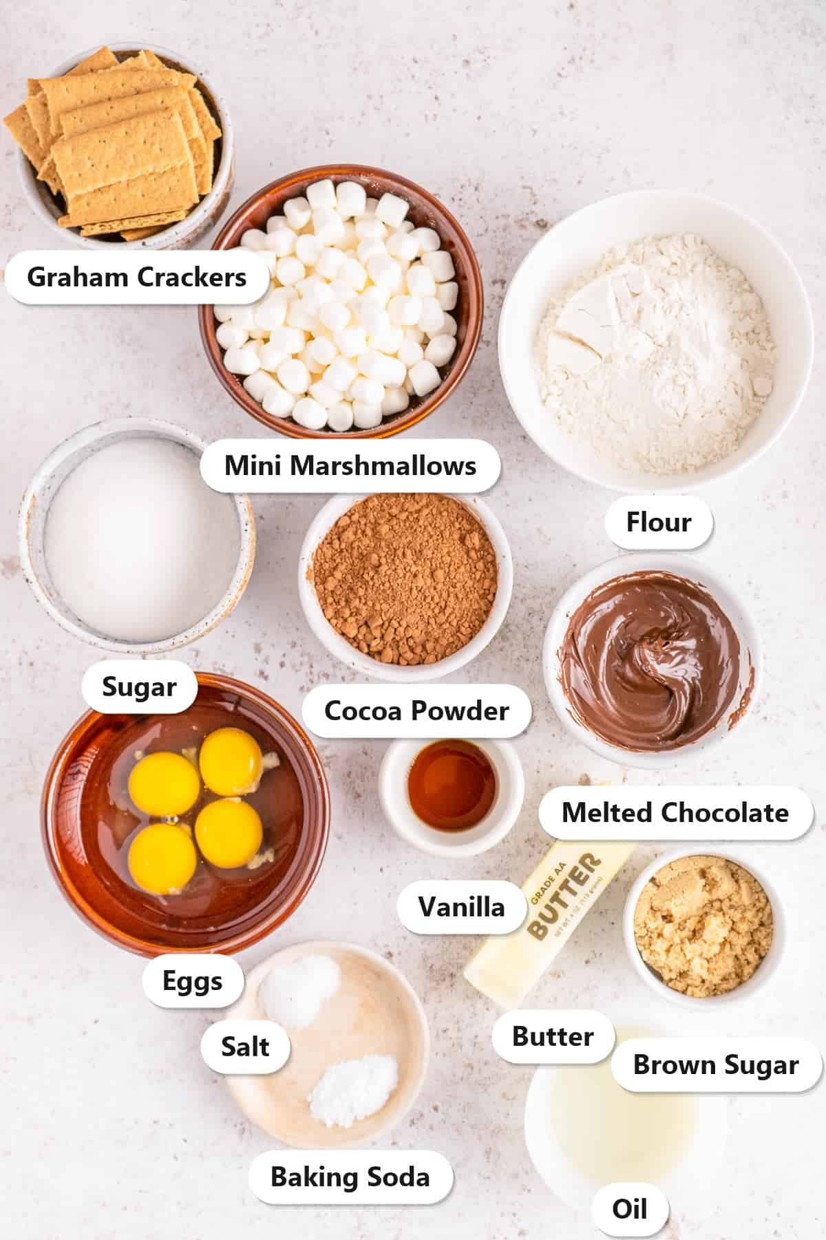 Ingredients to make smores brownies: graham crackers, mini marshmallows, flour, sugar, brown sugar, cocoa powder, melted chocolate, eggs, butter, oil, vanilla, salt, and baking soda.
