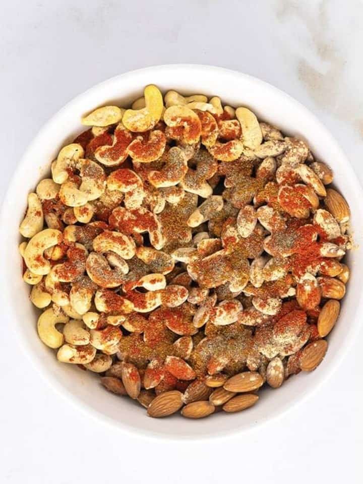 Raw almonds and cashews in a white bowl topped with olive oil and spices for spiced nuts.