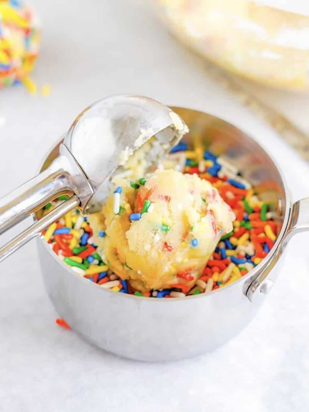 A cookie dough ball being rolled in a measuring cup full of rainbow sprinkles.