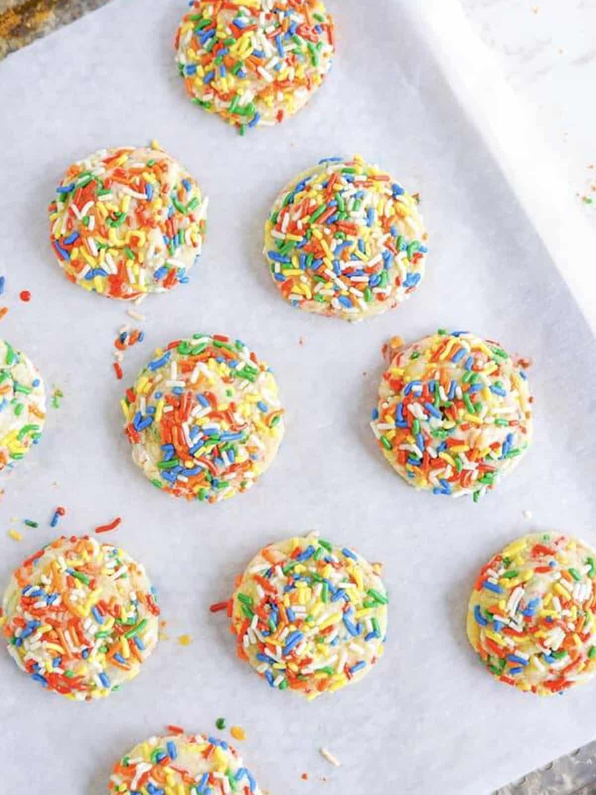 Baked Sprinkle Cookies covered in rainbow sprinkles on a parchment lined baking sheet.