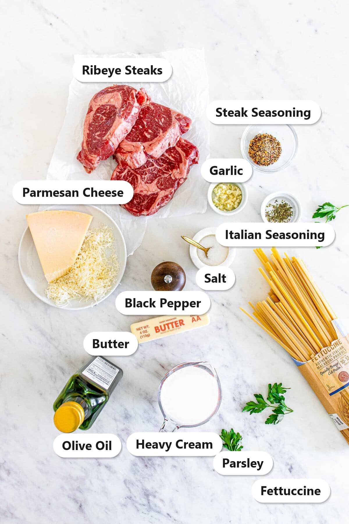 Labeled ingredients to make Steak Alfredo, ribeye steaks, fettuccine, Parmesan cheese, heavy cream, butter, garlic, and seasonings.