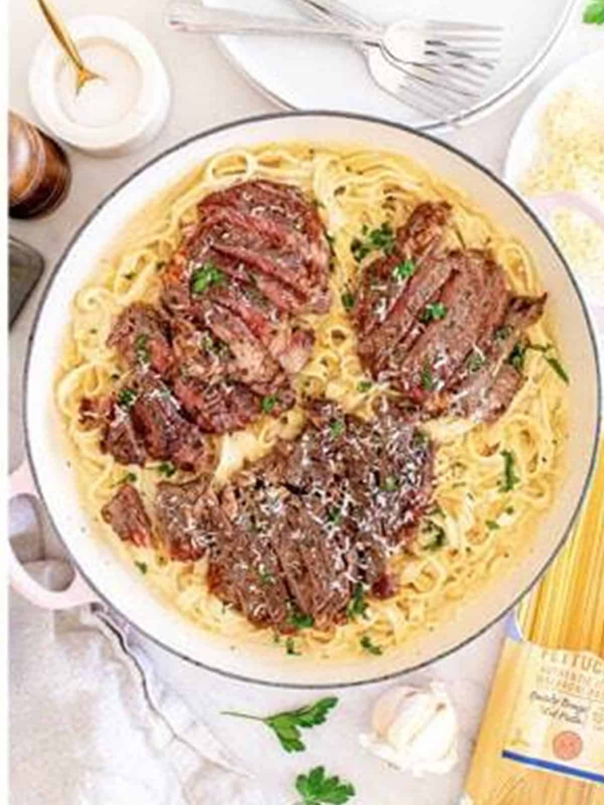 Sliced ribeye steak arranged over fettuccine alfredo in the skillet with parsley.