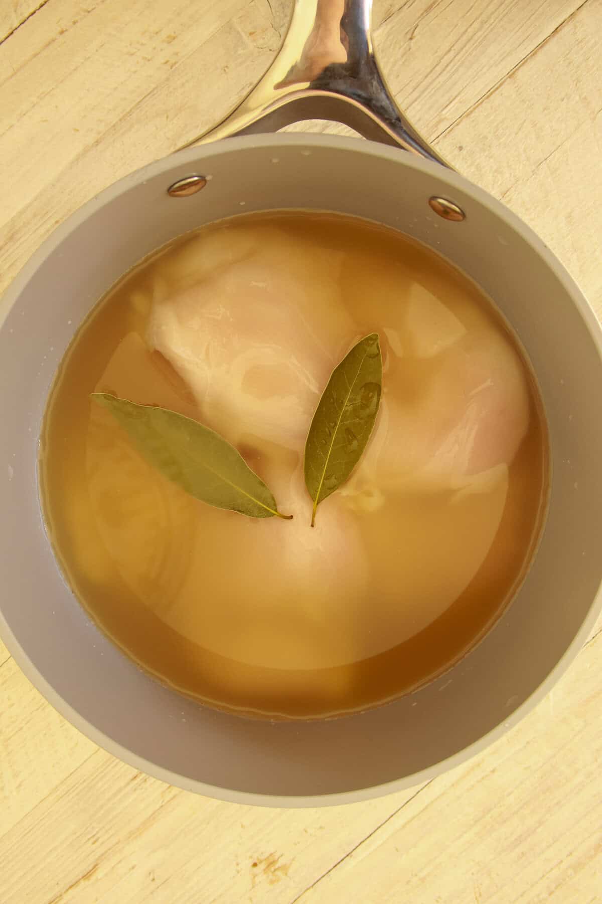 A split chicken breast simmering with bay leaves in chicken stock in a pot.