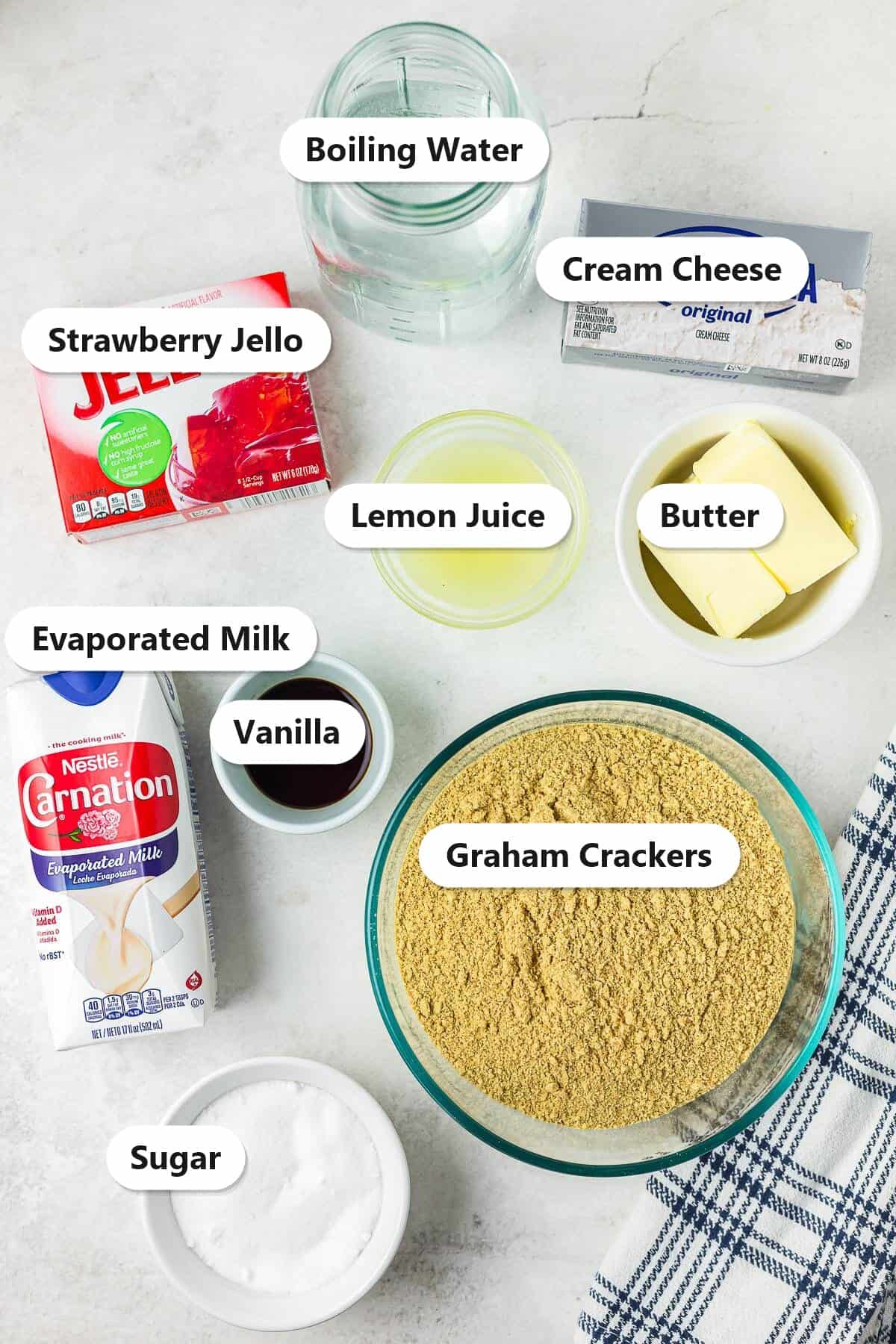 Labeled ingredients to make no bake strawberry icebox cake including strawberry jello, evaporated milk, cream cheese, lemon juice, vanilla, butter, graham crackers, and sugar.
