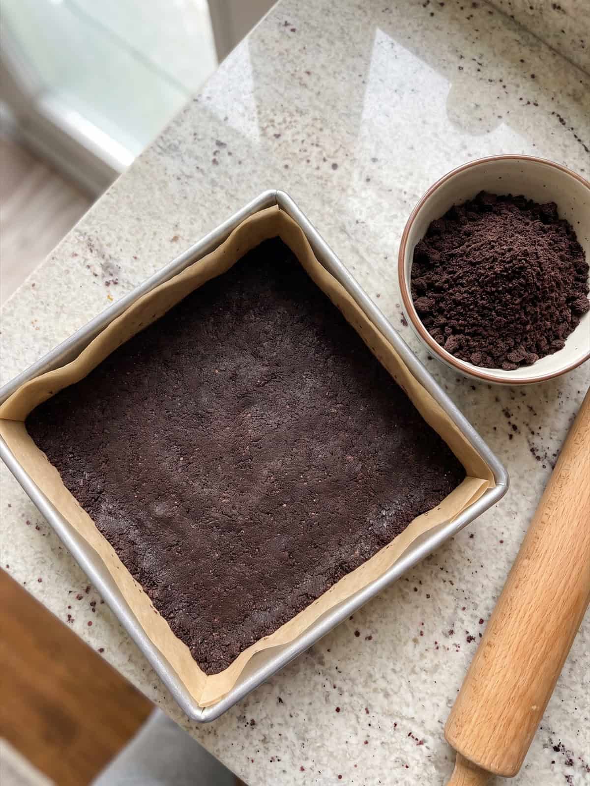 A pressed chocolate cookie crumb crust in a parchment lined pan for strawberry icebox cake bars.
