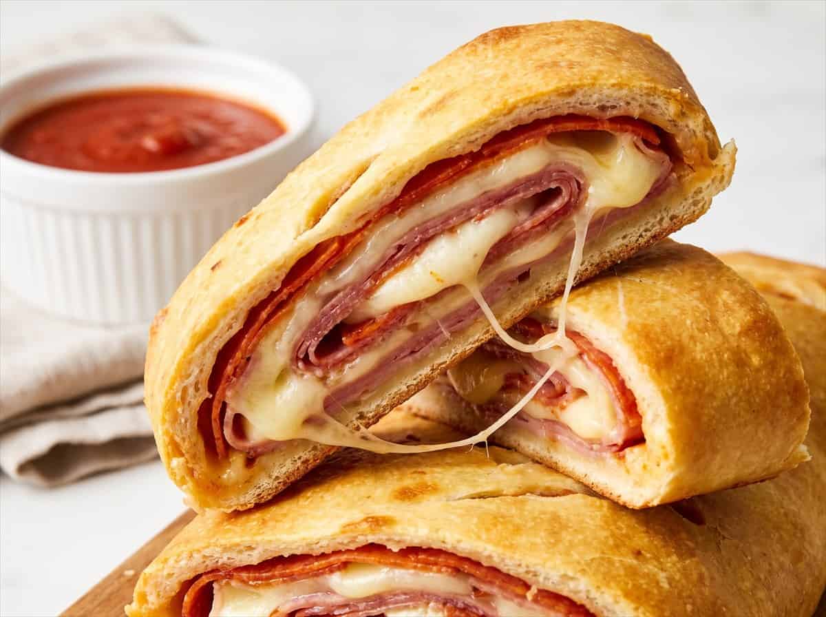Stromboli slice with a melted mozzarella cheese pull and marinara sauce in the background.