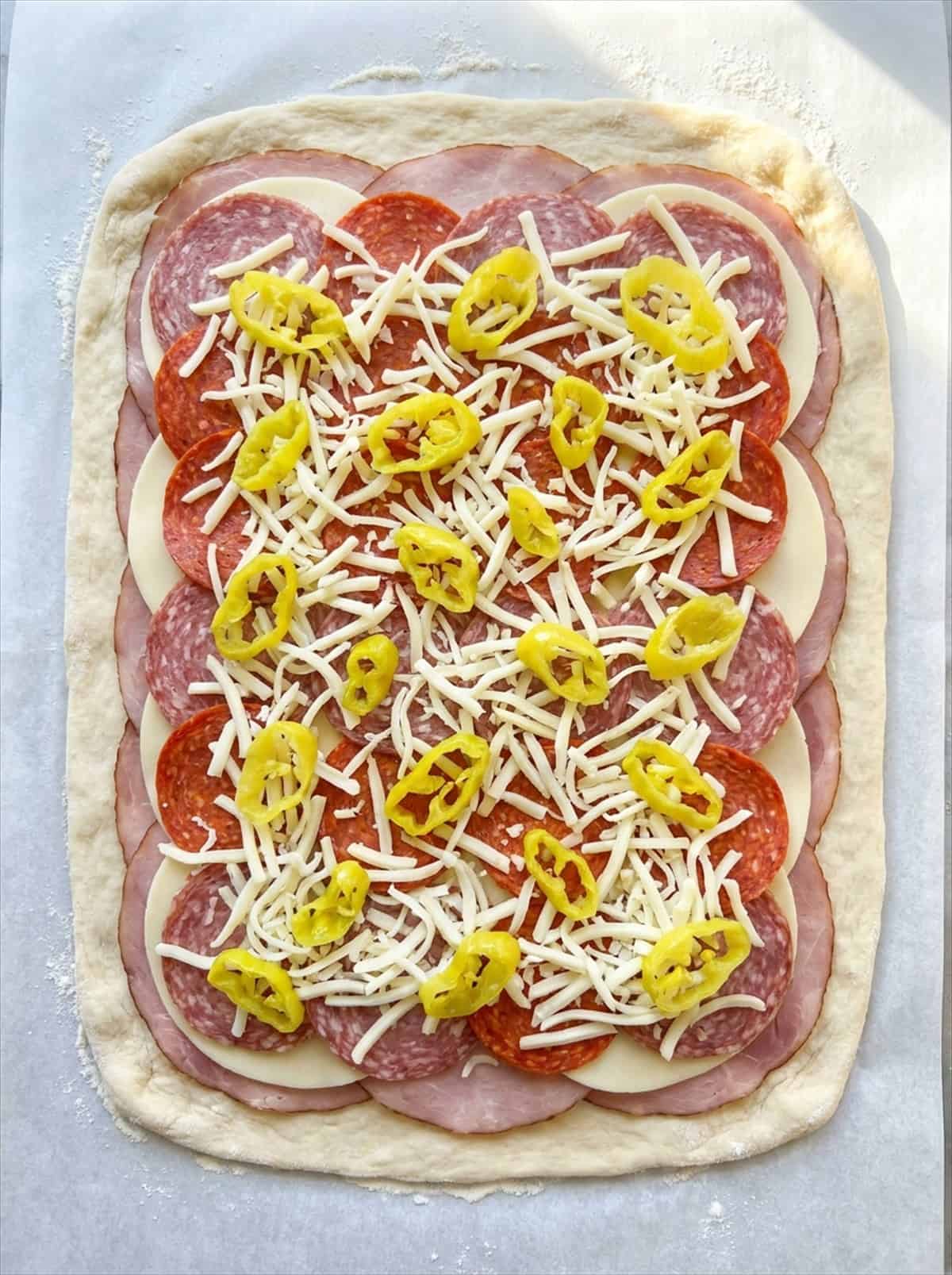 Rolled out pizza dough layered with ham, salami, pepperoni, provolone, mozzarella, and pepperoncini.
