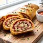 Baked stromboli sliced into pinwheel pieces on a cutting board with a bowl of marinara dipping sauce.