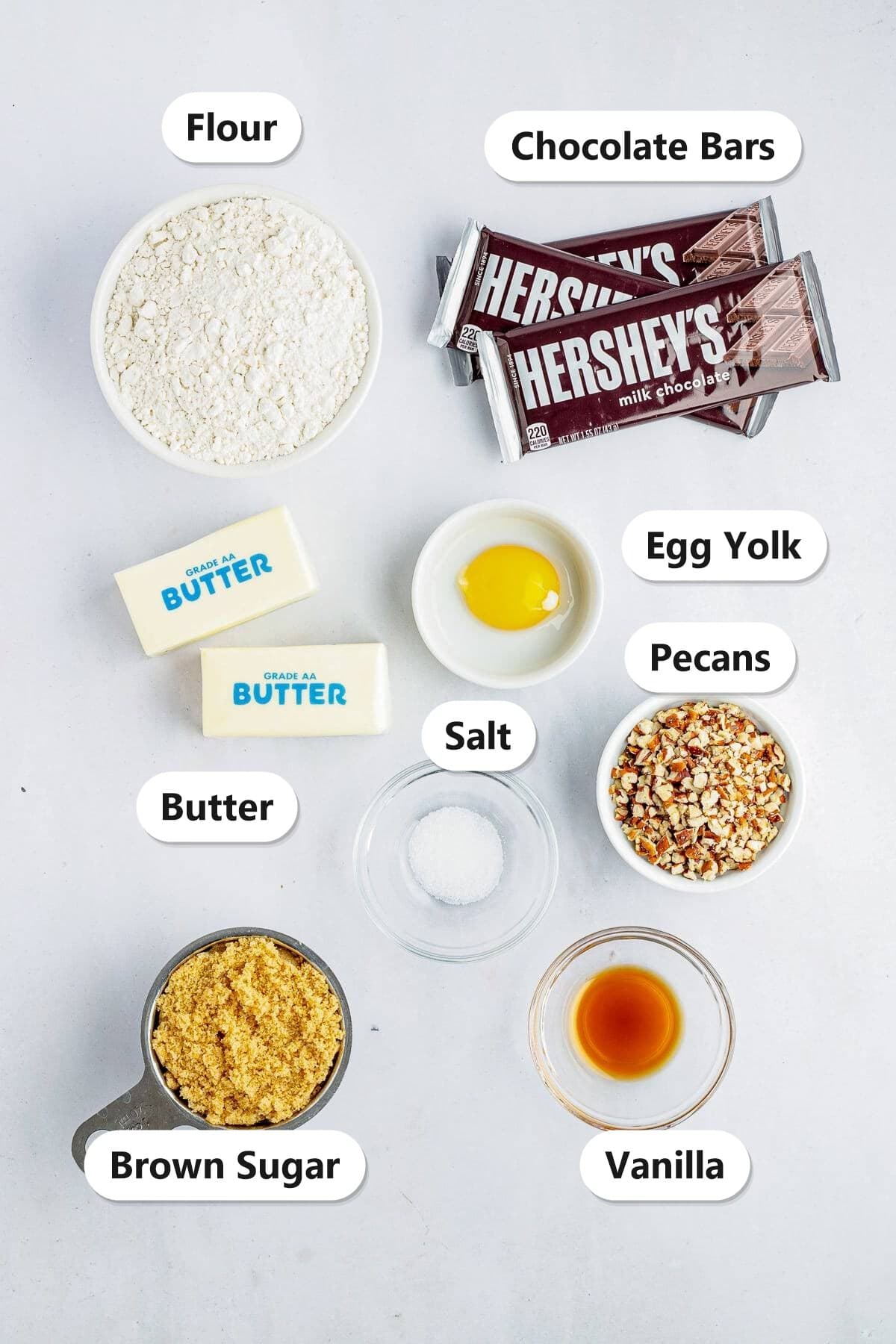 Ingredients to make toffee bars labeled on a white surface: flour, chocolate bars, butter, egg yolk, salt, pecans, brown sugar, and vanilla.