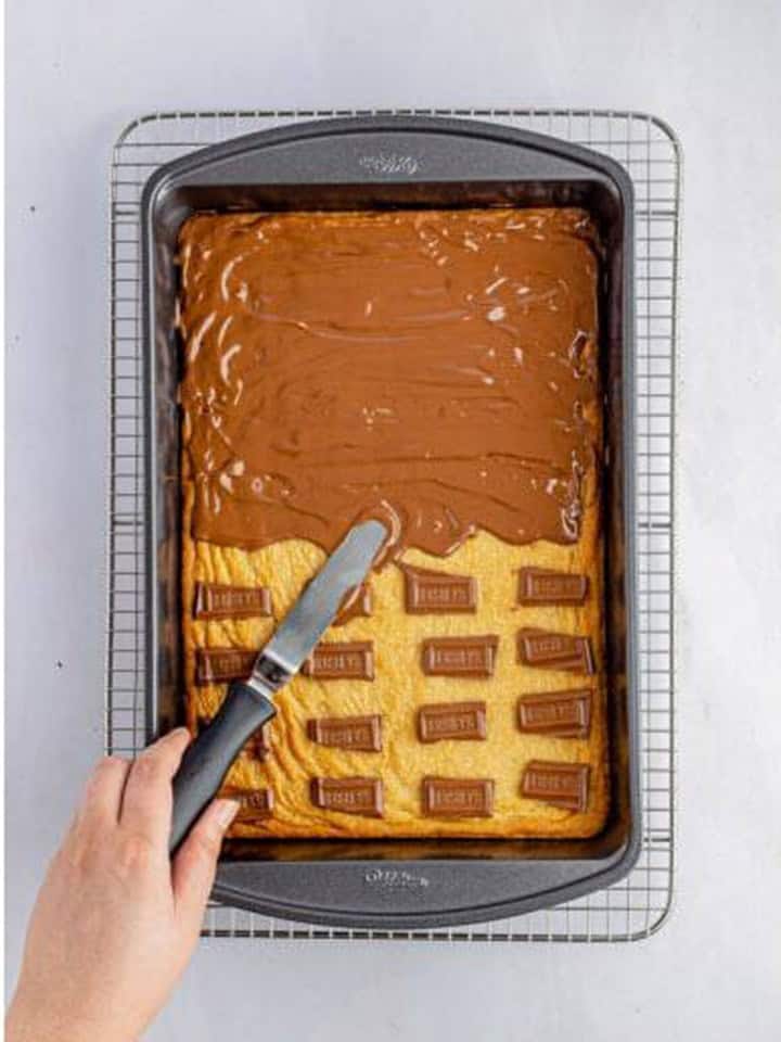 Melted chocolate bars being smoothed over the hot shortbread base with an offset spatula.