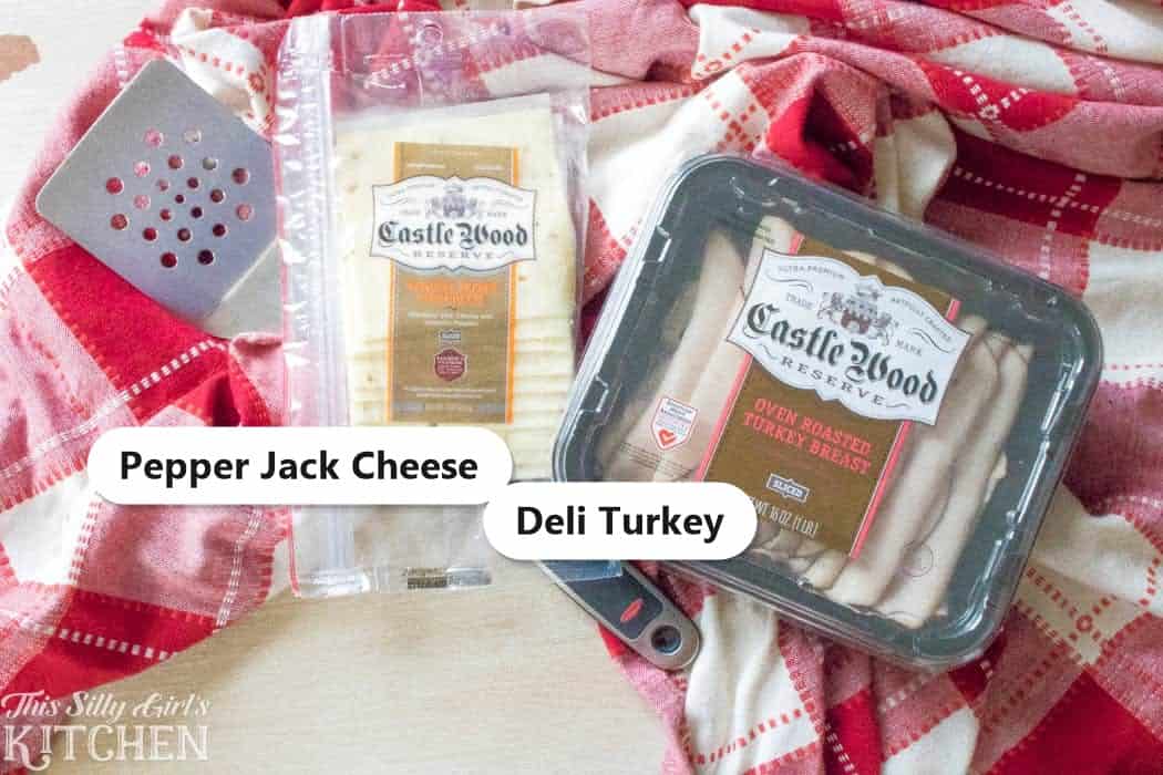 The simple ingredients for turkey sliders, deli turkey and pepper jack cheese, labeled on a red checkered cloth.