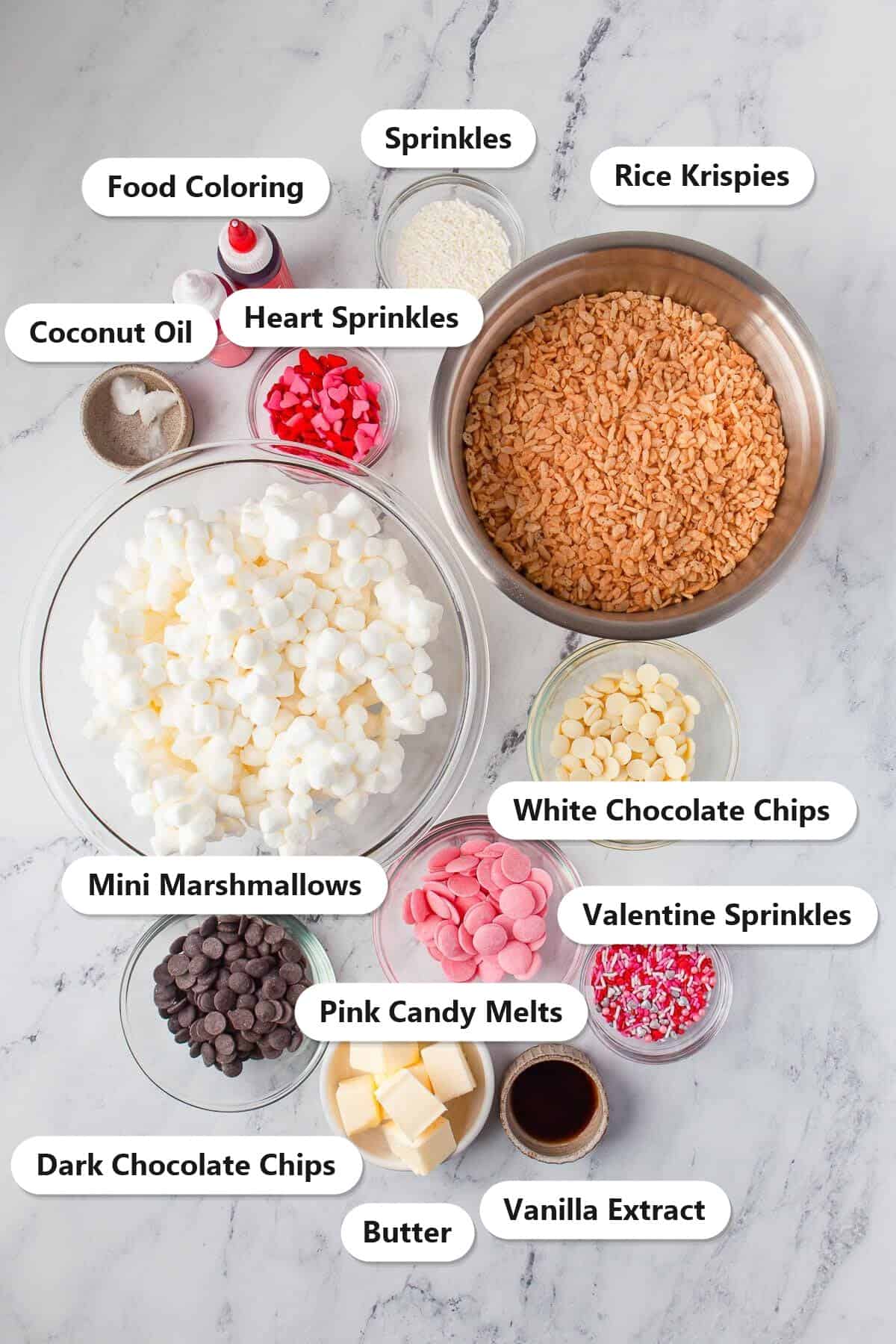Labeled ingredients for Valentine rice krispie treats including Rice Krispies, mini marshmallows, butter, vanilla extract, white chocolate chips, pink candy melts, dark chocolate chips, coconut oil, food coloring, and sprinkles.