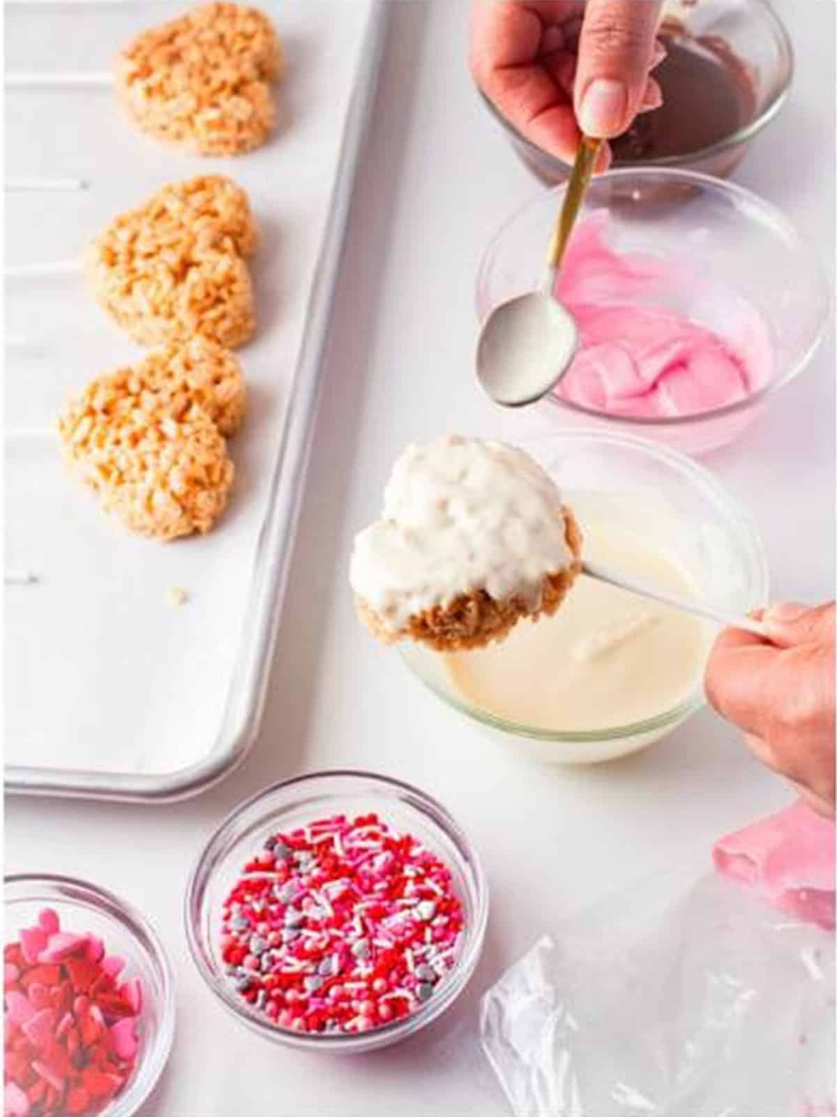 Rice krispie treat heart on a stick being dipped into melted white chocolate with a spoon.