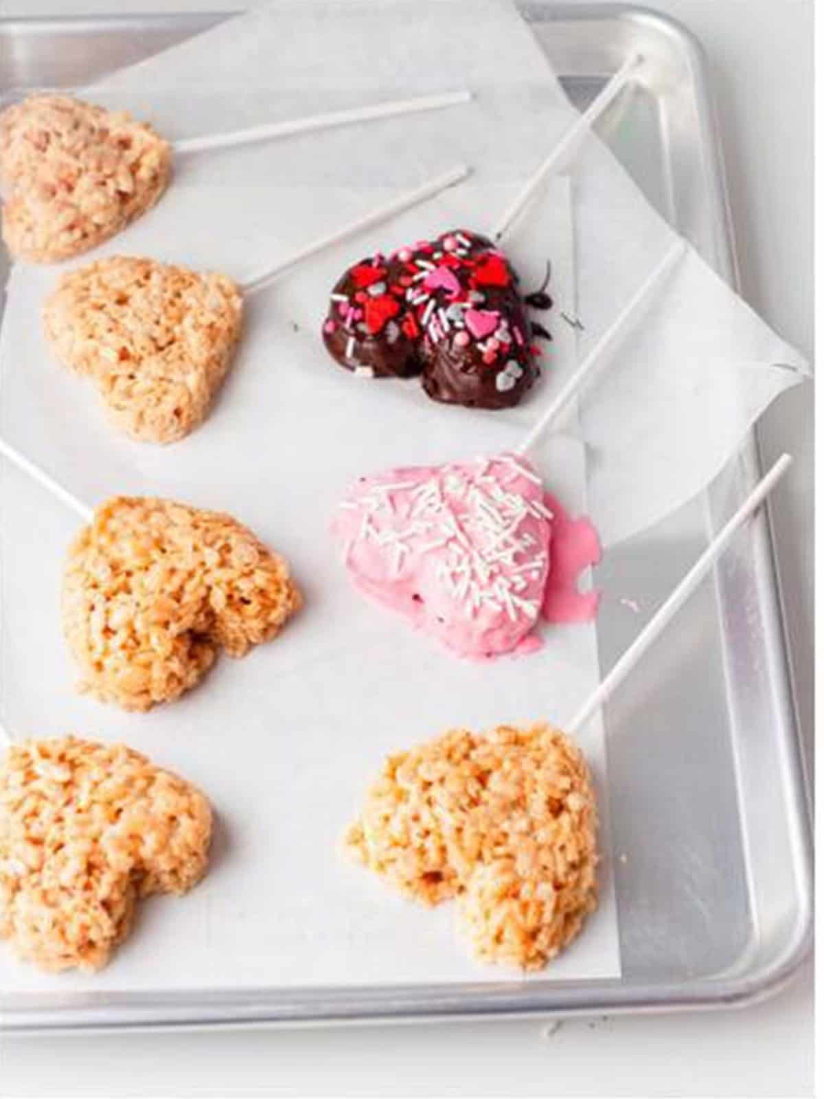 Chocolate dipped Valentine rice krispie treat hearts decorated with sprinkles setting on parchment paper.