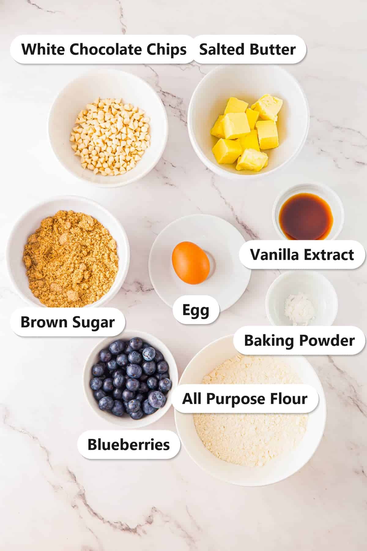 Labeled ingredients for white chocolate blondies: white chocolate chips, salted butter, brown sugar, egg, vanilla extract, baking powder, blueberries, and flour.