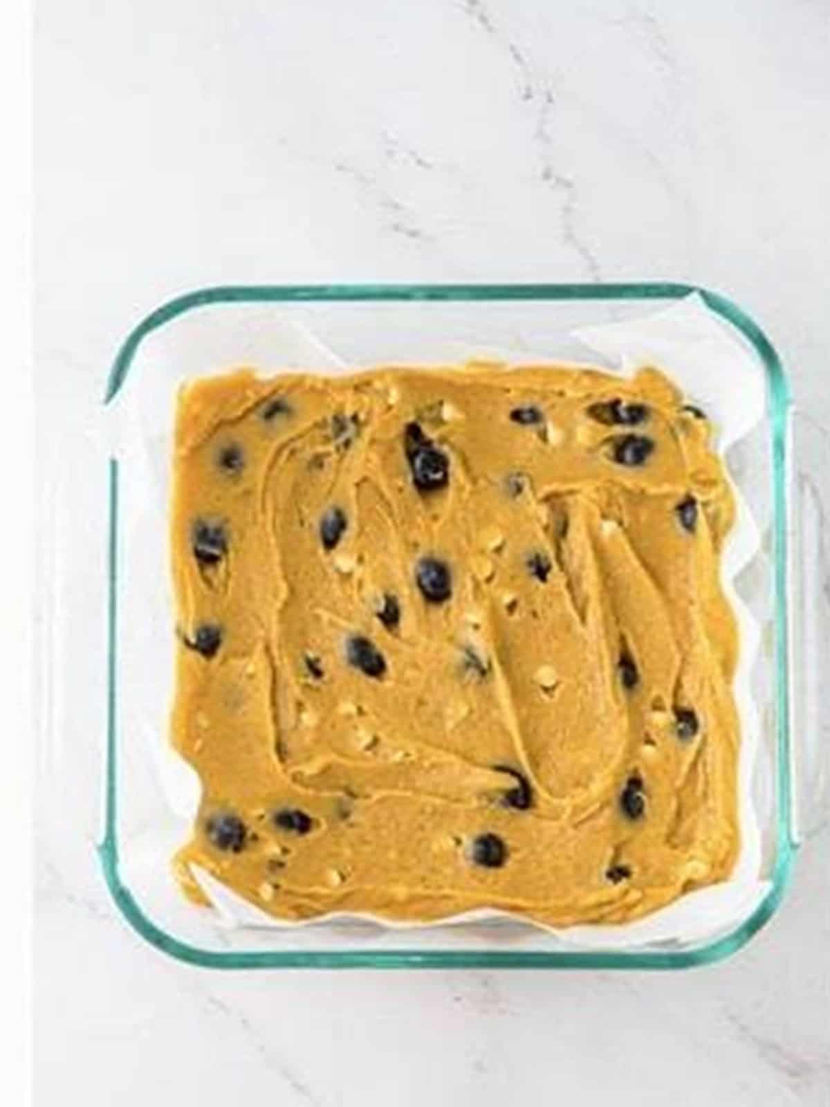 Blueberry white chocolate blondie batter spread evenly in a parchment lined glass baking pan.