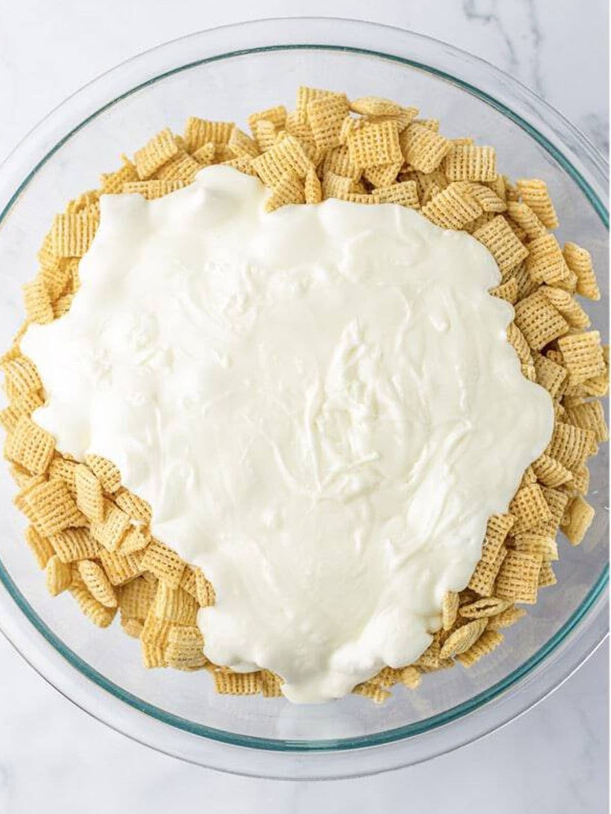 Melted white almond bark poured over rice chex cereal in a glass bowl for white chocolate chex mix.