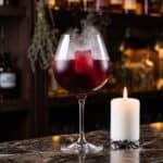 A Witches Brew Halloween cocktail in a stemmed goblet with red cranberry juice ice cubes and spooky smoke drifting over the rim.