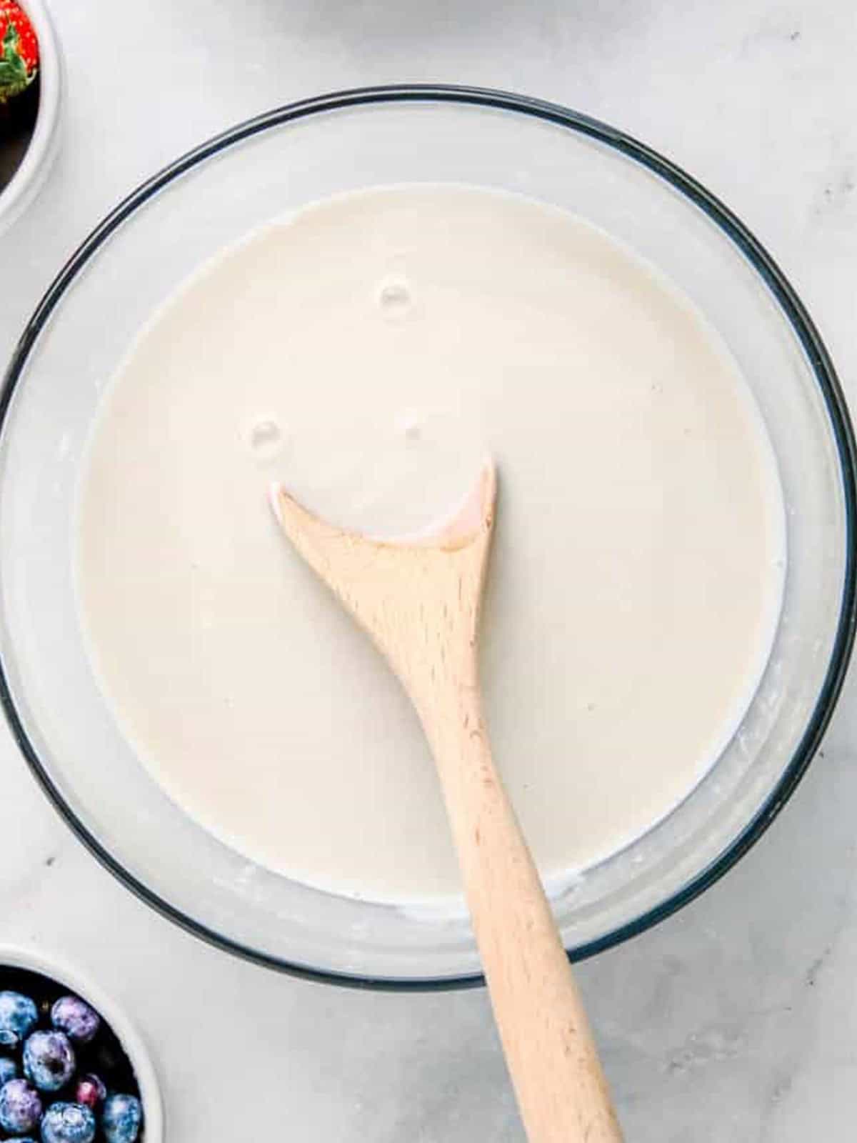 Yogurt, milk, maple syrup, and vanilla stirred together with a wooden spoon in a glass bowl.