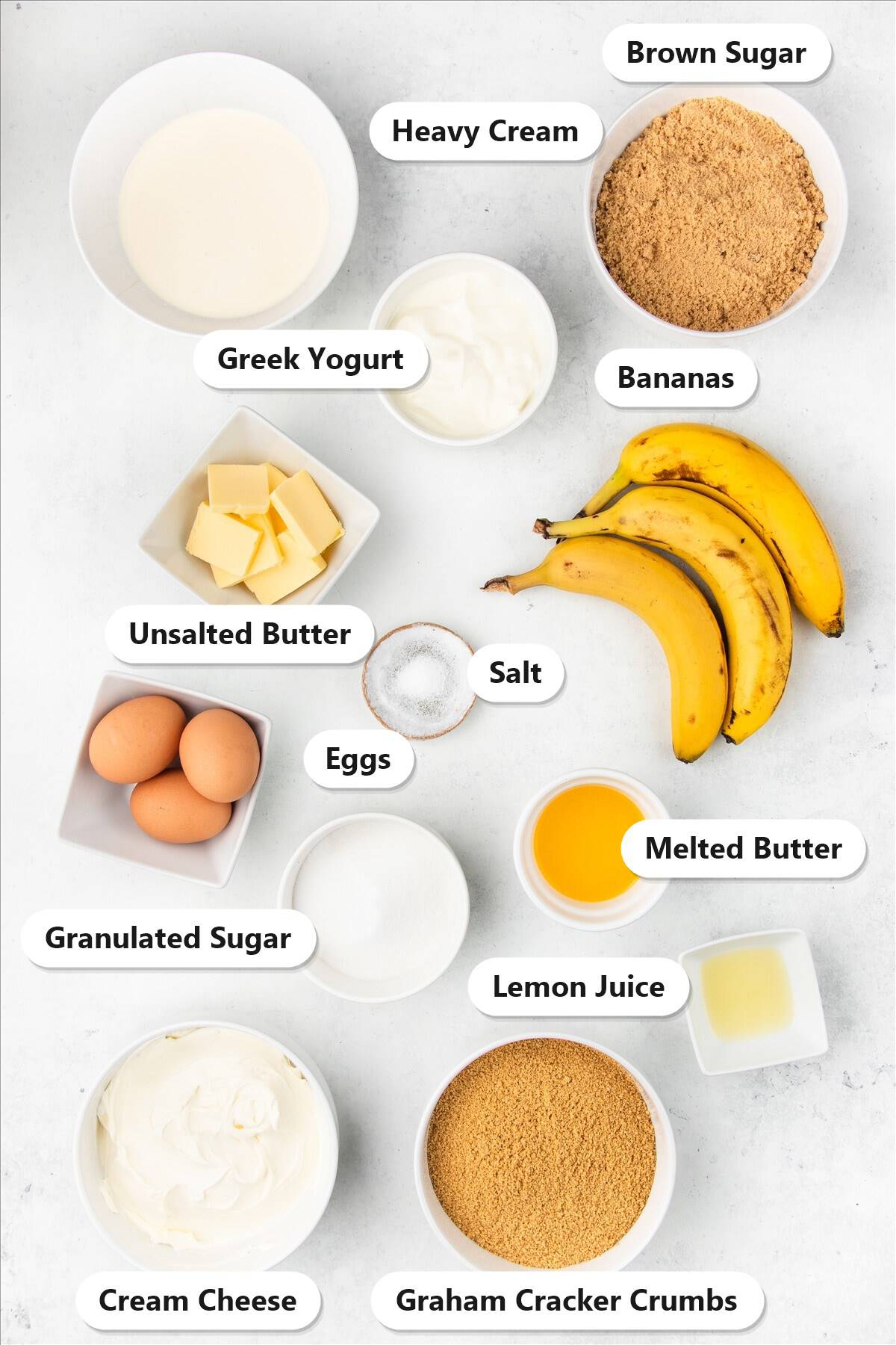 Labeled ingredients for banoffee cheesecake including graham cracker crumbs, butter, ripe bananas, cream cheese, Greek yogurt, eggs, sugar, brown sugar and heavy cream.