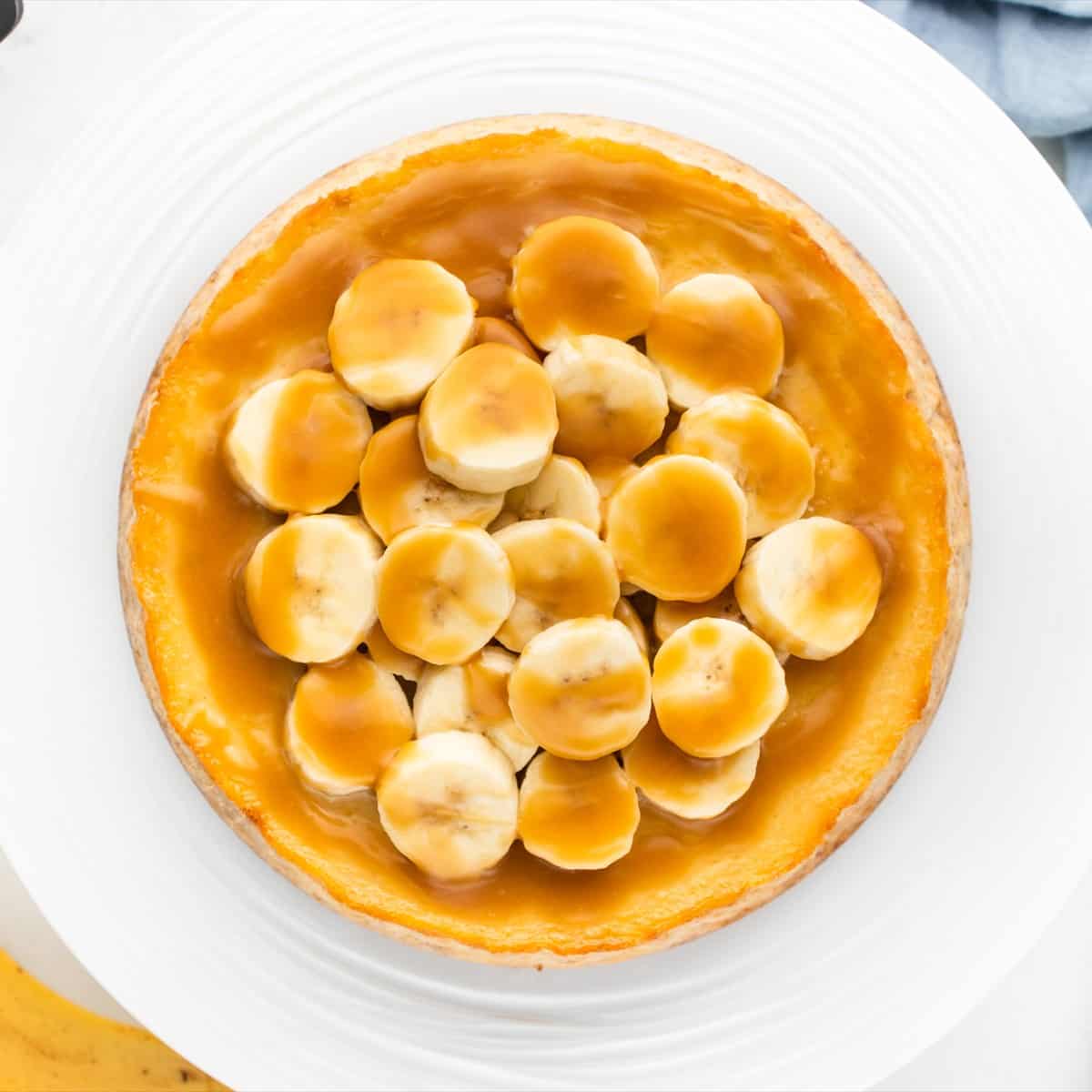 Banoffee cheesecake on a cake stand with toffee sauce poured over the top and banana slices on the edge.