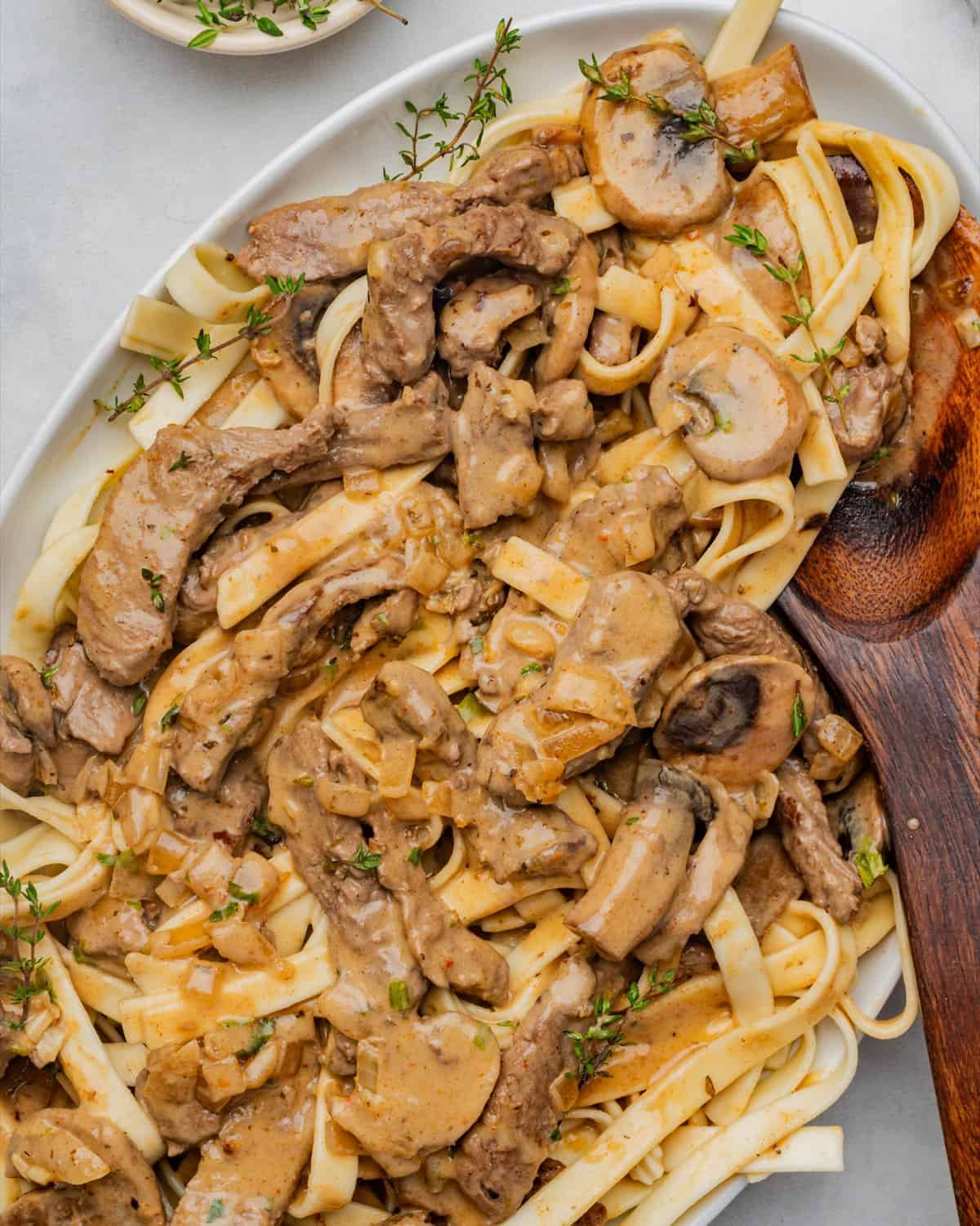 Beef stroganoff served on a large oval platter over egg noodles with a wooden spoon and fresh thyme.