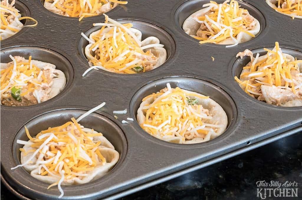 Shredded cheddar cheese sprinkled over chicken enchilada bites in a muffin tin before baking.