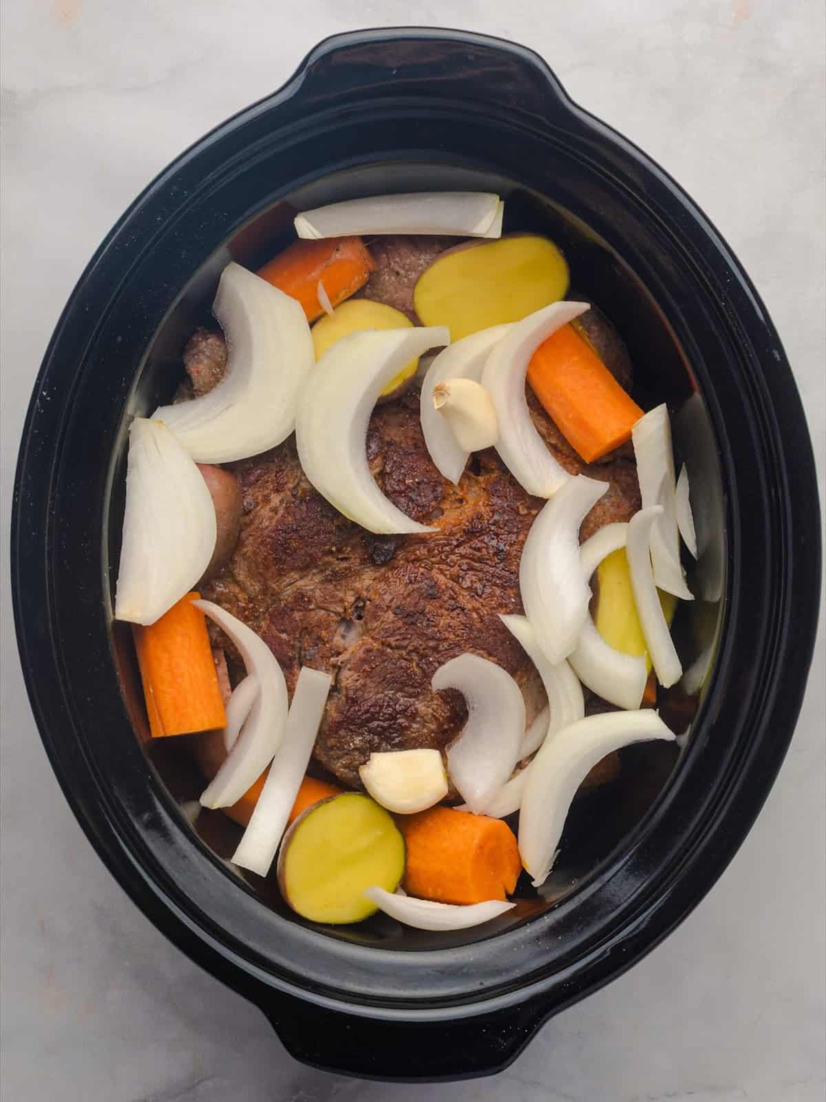 Seared chuck roast layered over carrots, potatoes and onions in a slow cooker.
