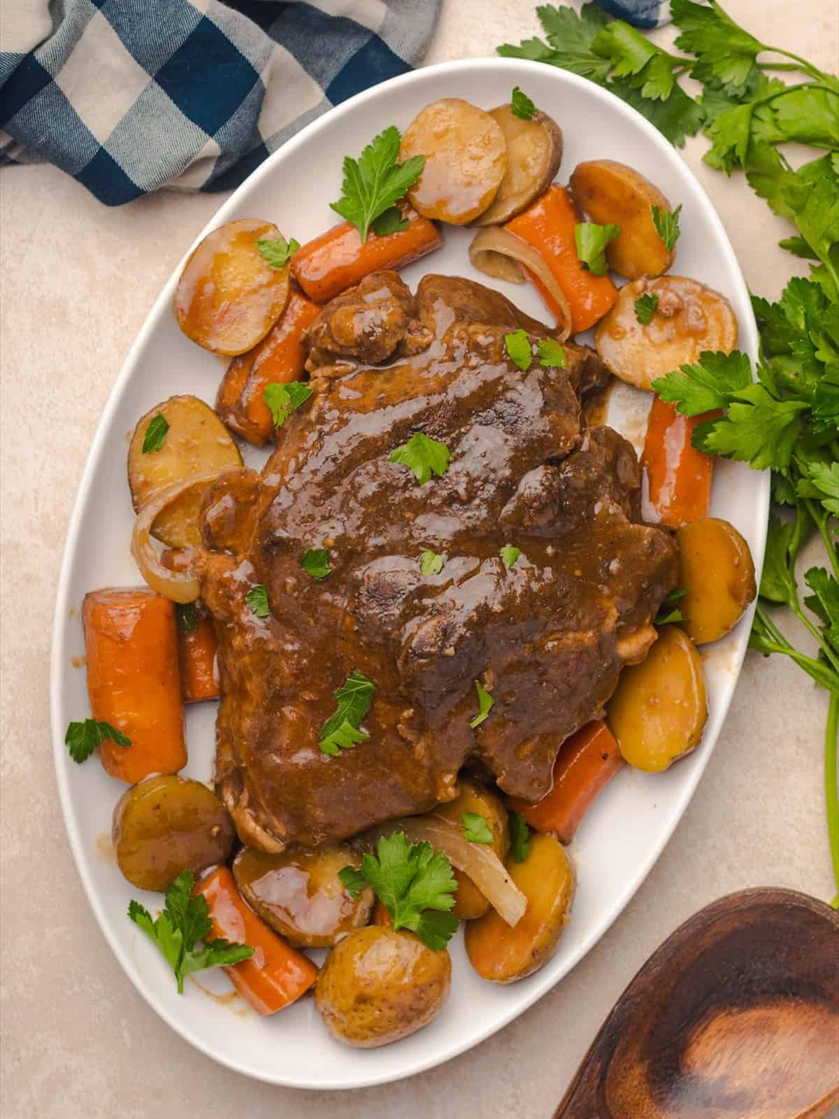 Crock pot pot roast served on a white oval platter with carrots, potatoes and fresh parsley.