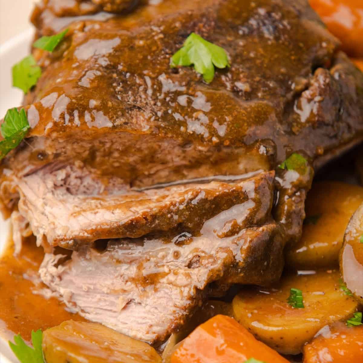 Sliced crock pot pot roast on a white platter with carrots and baby potatoes and brown gravy.