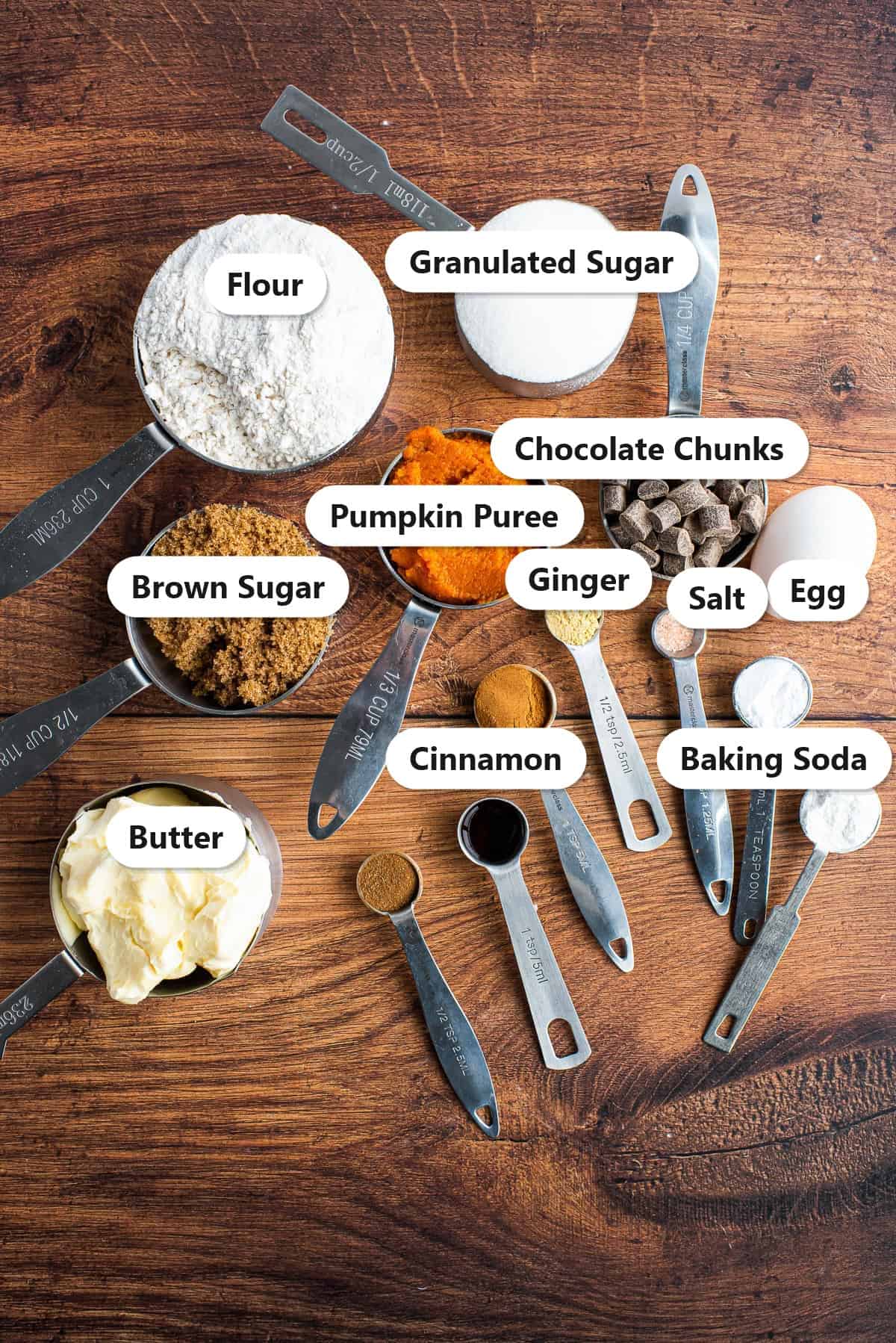 Labeled ingredients for pumpkin chocolate chip cookies including flour, granulated and brown sugar, pumpkin puree, chocolate chunks, egg, butter, cinnamon, ginger, salt, and baking soda.