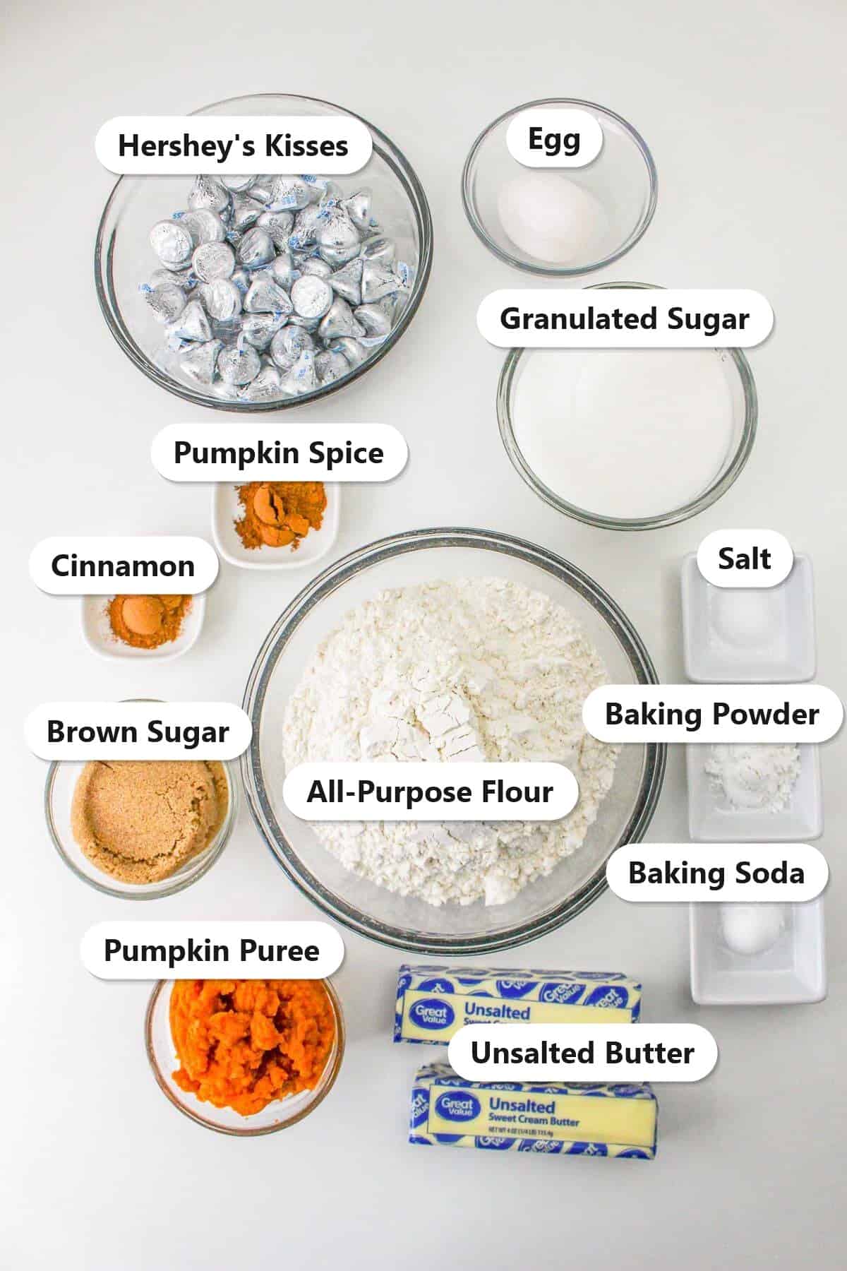 Labeled ingredients for pumpkin cookies including all-purpose flour, pumpkin puree, granulated and brown sugar, unsalted butter, egg, pumpkin spice, cinnamon, baking powder, baking soda, salt, and Hersheys Kisses.