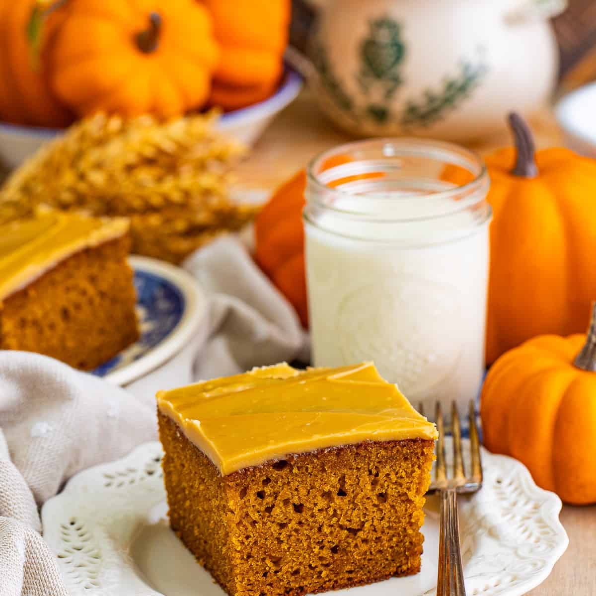 A square slice of pumpkin spice cake with brown sugar frosting on a white plate with a glass of milk and mini pumpkins.