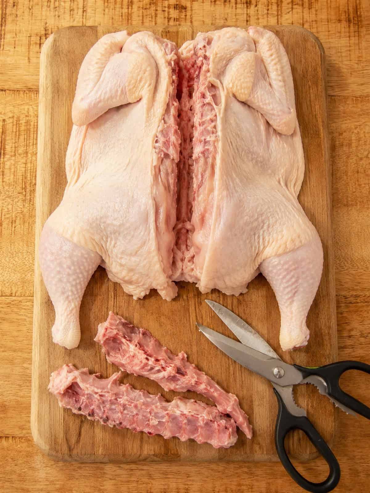 A whole raw chicken breast side down with the backbone removed and kitchen shears on a wooden cutting board.
