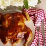 A whole smoked spatchcock chicken glazed with barbecue sauce on a white plate with a red gingham towel.