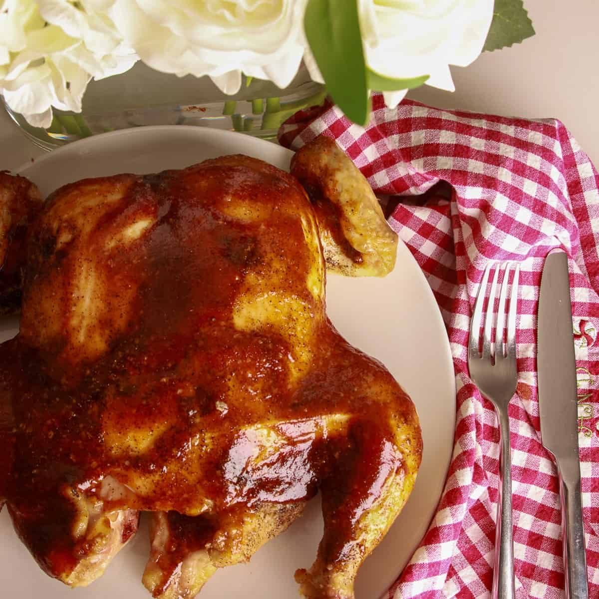 A whole smoked spatchcock chicken glazed with barbecue sauce on a white plate with a red gingham towel.