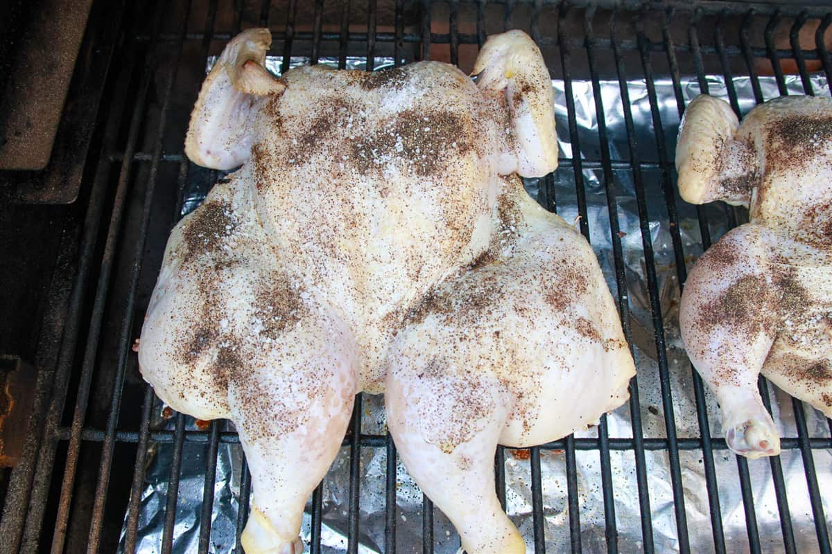 A seasoned raw spatchcock chicken positioned skin side up on the smoker grill grates.