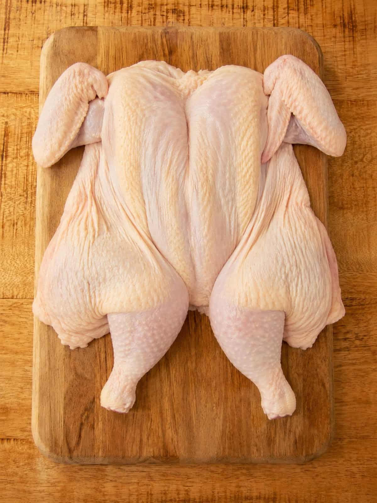 A raw spatchcock chicken pressed flat breast side up on a wooden cutting board.