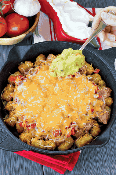 Guacamole being layered over melted cheese and tater tots for Totchos.