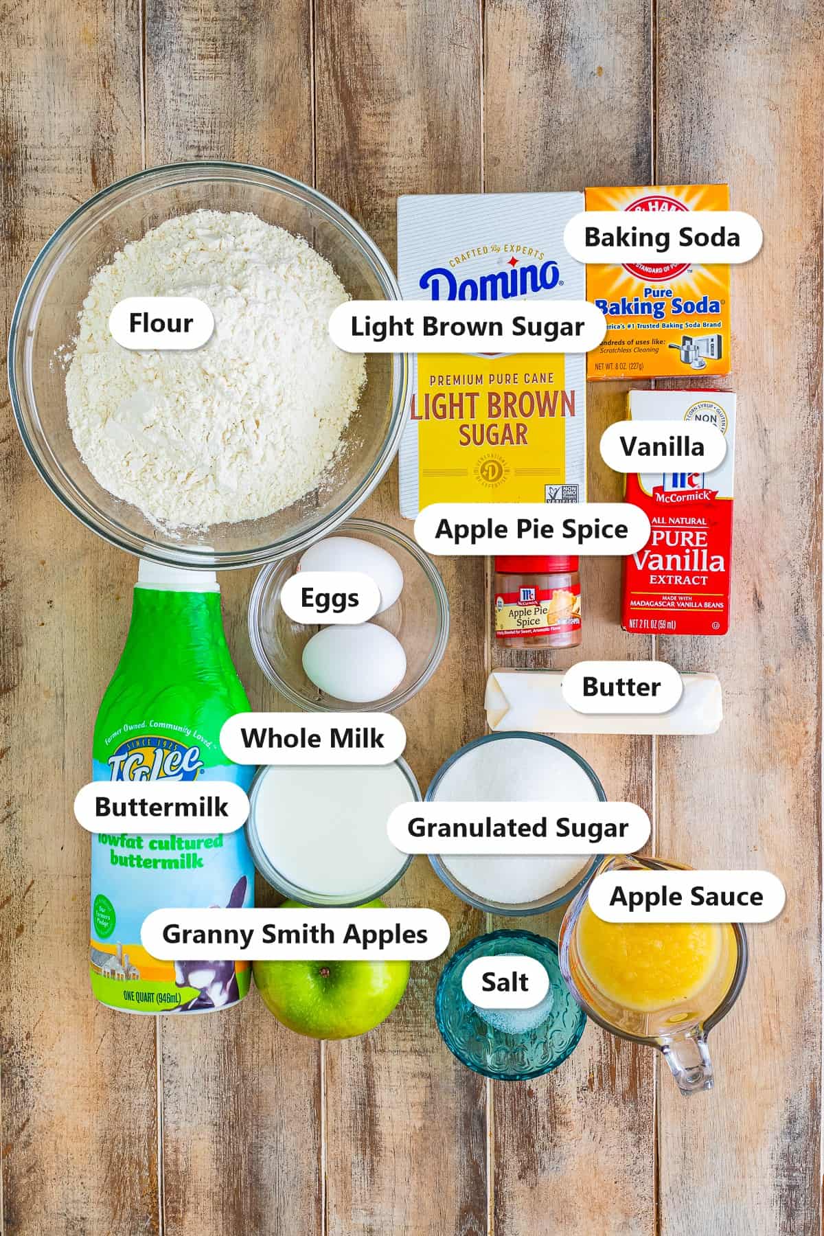 Labeled ingredients for apple spice cake including flour, apple pie spice, baking soda, salt, eggs, buttermilk, whole milk, granulated sugar, light brown sugar, butter, apple sauce and Granny Smith apples.