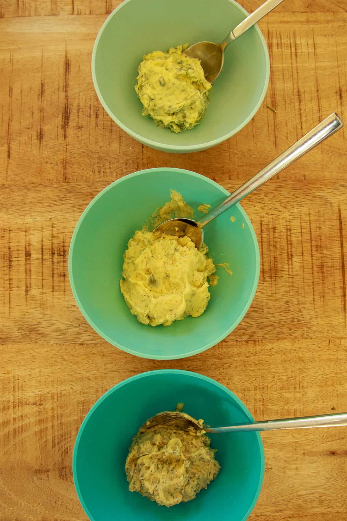 Three bowls of compound butter mixed with herbs, olives, and scallion dill before rolling into logs.