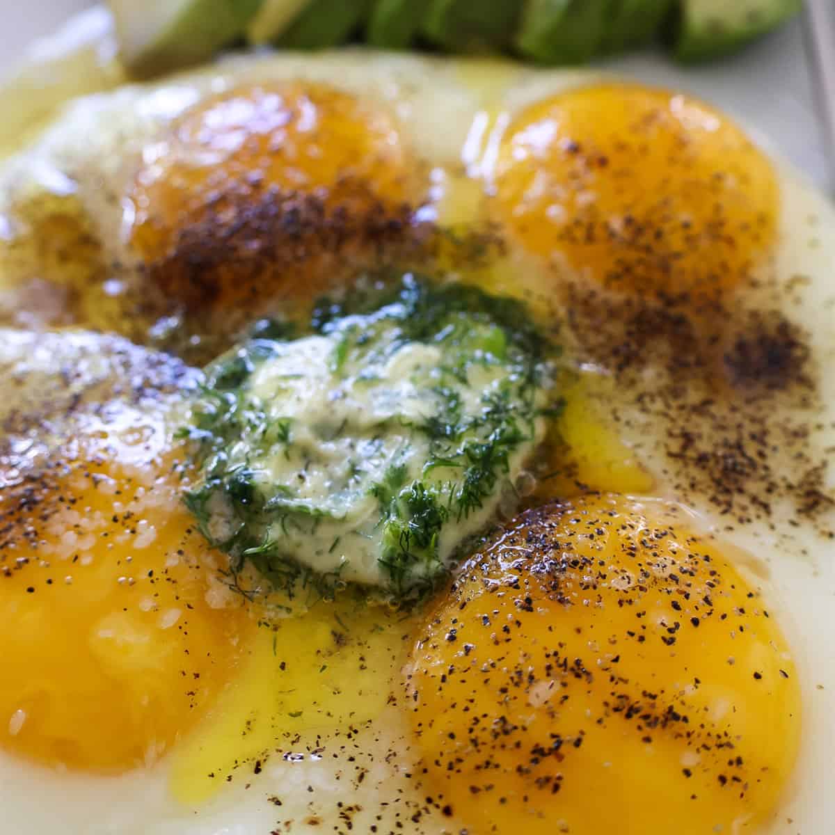 Scallion dill compound butter melting over fried eggs with sliced avocado on a white plate.