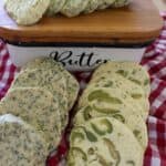 Sliced compound butter logs with herb garlic and green olive flavors next to a butter dish on a checkered towel.